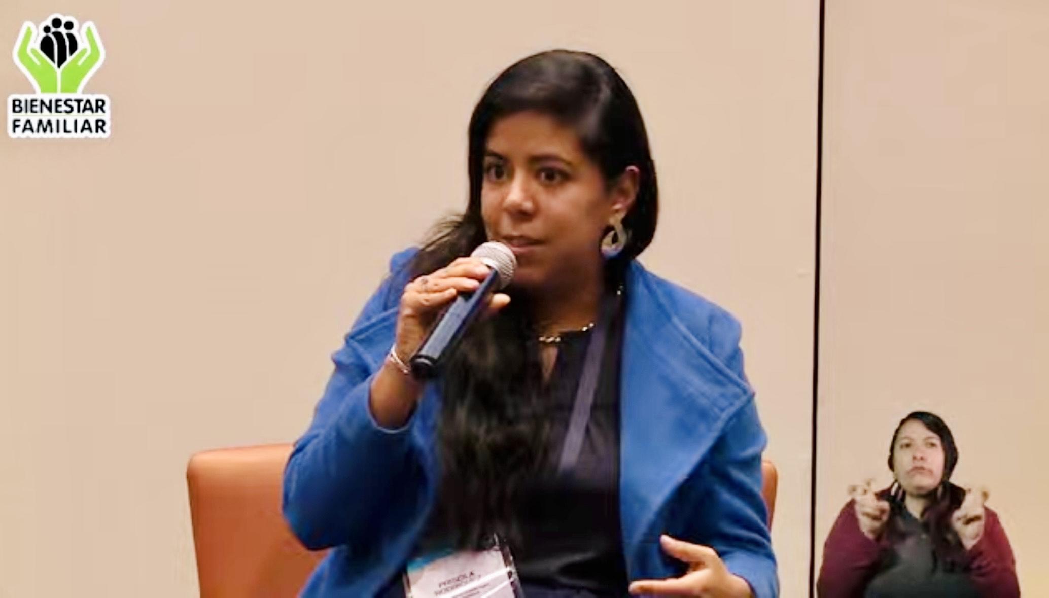 Priscila Rodriguez is seated speaking into a microphone, a sign language interpreter is in view to her right