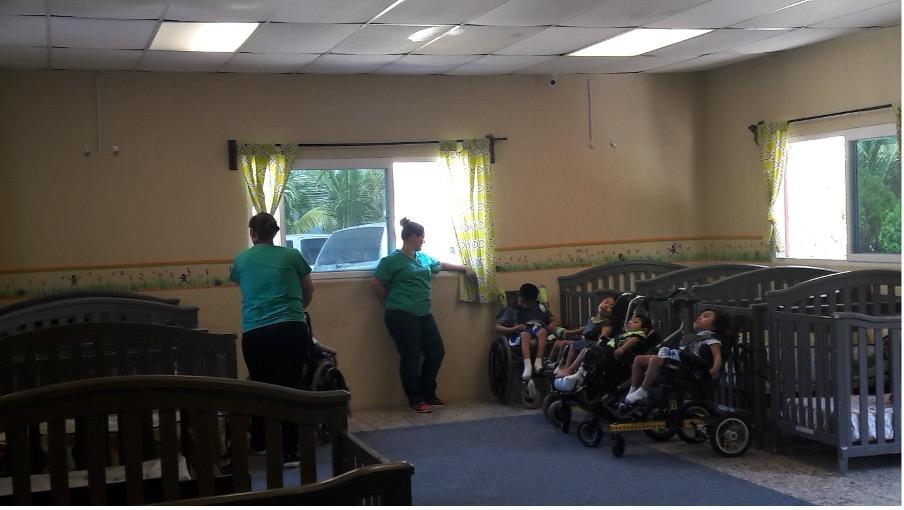 A group of children in wheelchairs in a room full of cribs